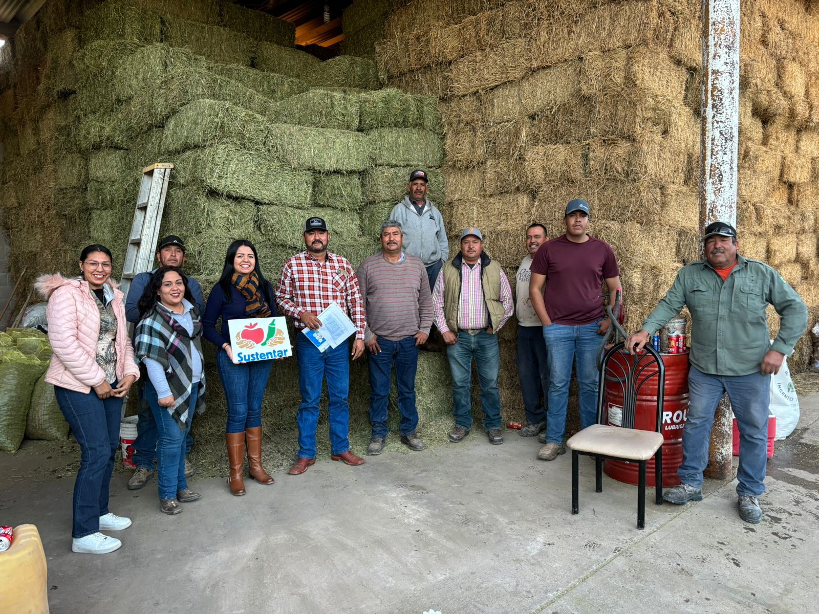 Group photo of people standing in front of a large stack of hay bales. One of them is holding a Sustentar poster.